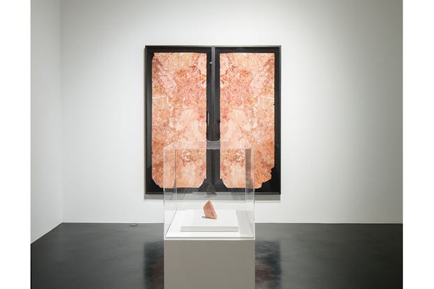 A peachy colored marbled rock sits above a white stand surrounded by glass. In the center behind it on the wall is a print of a black background with two prints similar in color and pattern to the rock divided in the middle.  