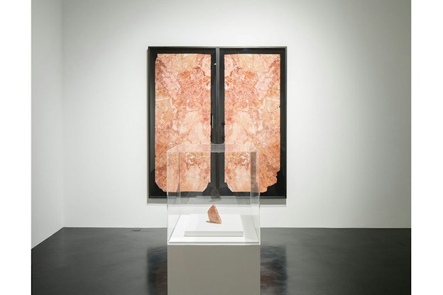 A peachy colored marbled rock sits above a white stand surrounded by glass. In the center behind it on the wall is a print of a black background with two prints similar in color and pattern to the rock divided in the middle.  