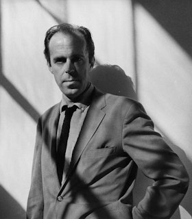 A black and white photograph of Earle Brown wearing a suit and standing in front of a bare wall. Diagonal striped shadows bisect his body. With his hand on his hip, he looks directly at the camera. 