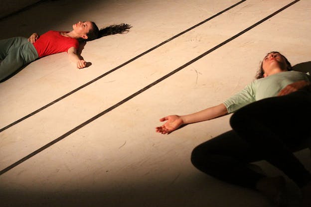 Slanted still of two persons lying on their backs on a dimly lit floor separated by two black lines. 