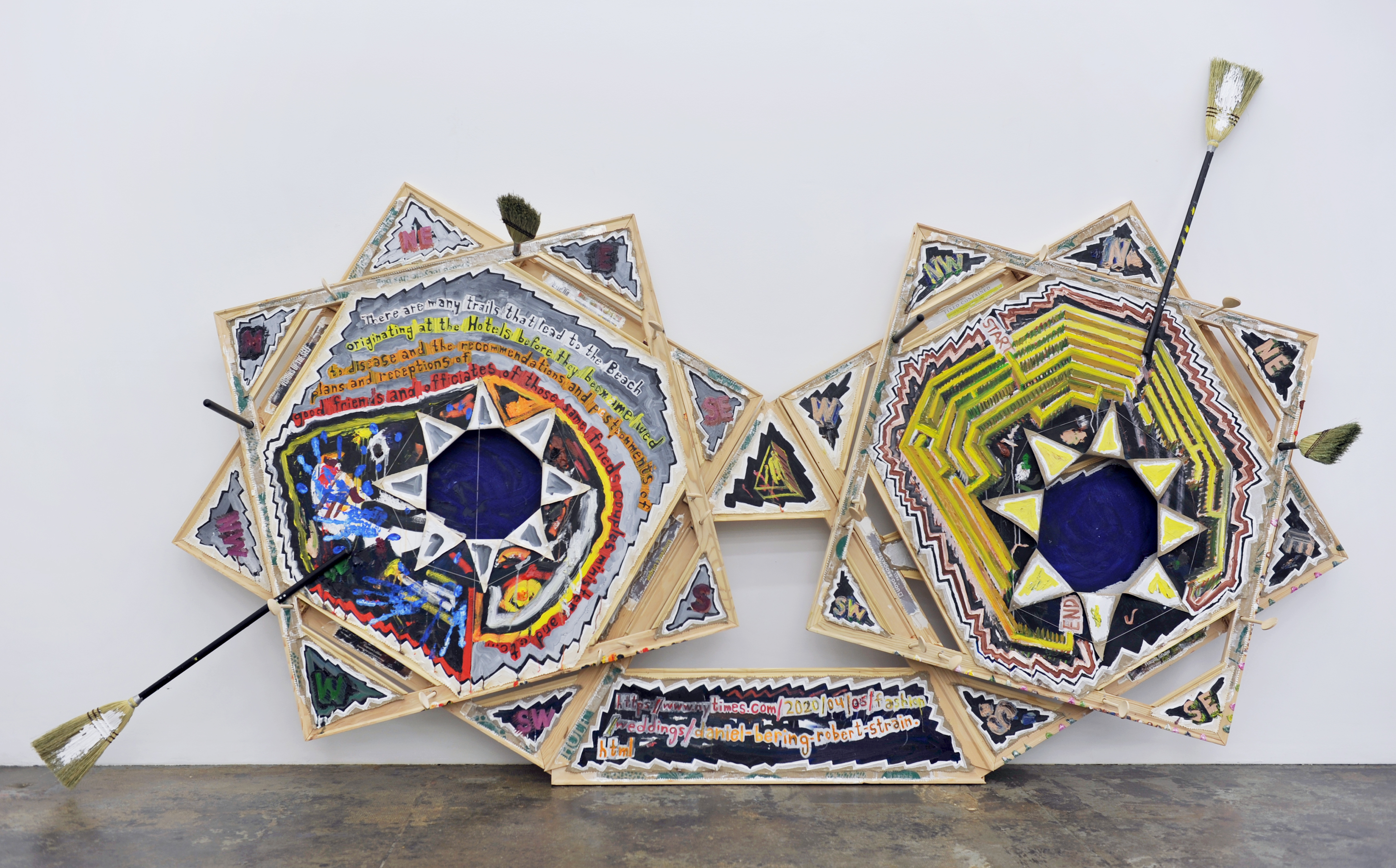 An installation view of freestanding abstract paintings on irregularly shaped canvases featuring triangles and bold lines. Brooms stick out of the canvas. Text on the painting reads: ‘There are many trails that lead to the Beach originating at the Hotels before they becmame [sic] wed/ to disease and the recommendations and postponments [sic] of plans and receptions of / good friends and officiates of those same frieds [sic] couples minister and etc…