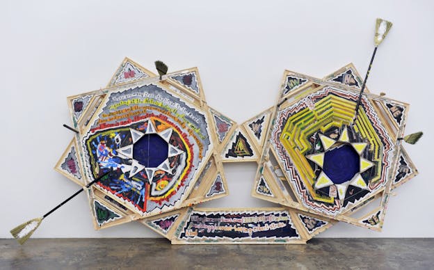 An installation view of freestanding abstract paintings on irregularly shaped canvases featuring triangles and bold lines. Brooms stick out of the canvas. Text on the painting reads: ‘There are many trails that lead to the Beach originating at the Hotels before they becmame [sic] wed/ to disease and the recommendations and postponments [sic] of plans and receptions of / good friends and officiates of those same frieds [sic] couples minister and etc…