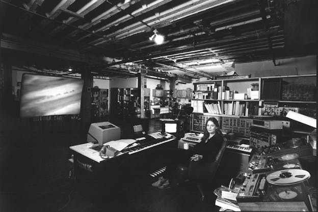 Black and white photograph of Laurie Spiegel with long straight hair in her studio seated in front of a computer and surrounded by three desks with pianos, radios, TV and other electronics between books. 