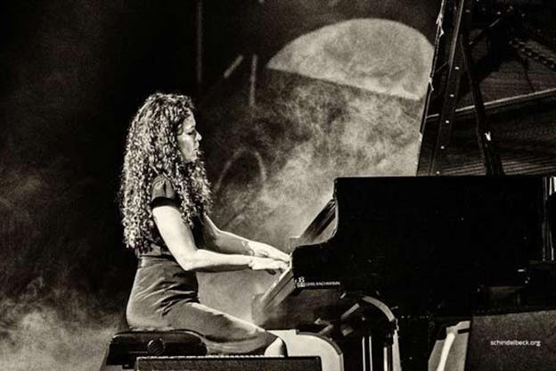 A black and white grainy photograph of a figure with long curly hair sitting profile to the viewer in front of a piano - playing. 