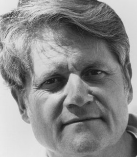 A close up black and white portrait of Gordon Mumma smiling slightly and looking towards the camera. He has short wavy hair. 