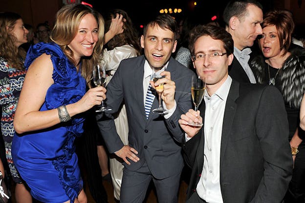 Flash photograph of a person in a cobalt blue minidress and two persons in suits holding up glasses of champagne and smiling in a room of people mingling.