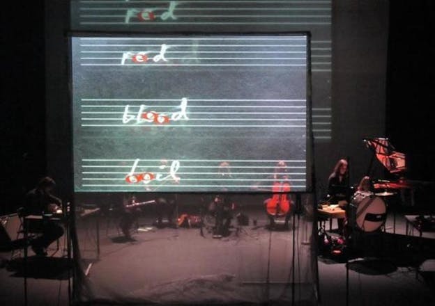 A small orchestra performs on a dimly-lit stage behind a clear screen projecting four rows of thin white lines and the words 