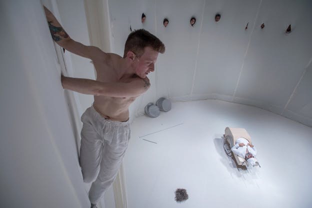 A close up photo of a shirtless person in white linen pants hanging from holes on a wall and looking towards a figure at the floor, dressed in white with black sunglasses laying inside a wooden box with two wheels and handles on each side. On the other side of the white room, aligned faces peak from holes carved on the wall.