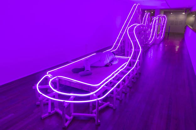 A structure similar to a fun park's train rails is lighted by purple neon lights on its outline. On the one side, closest to the viewer a figure lays down on a wooden board in its middle. 