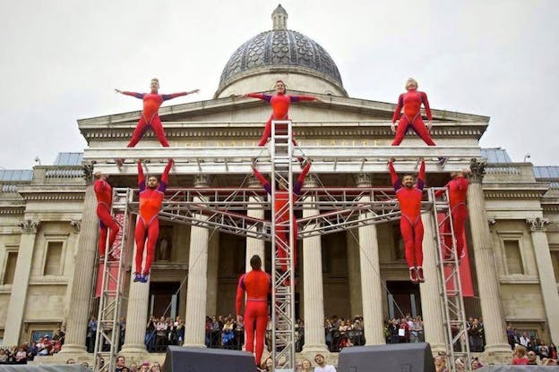 Performers dressed in red leotards surround a metallic structure in front of a historic building in various poses.