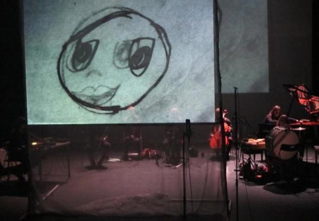 A small orchestra performs on a dimly-lit stage behind a screen projecting a sketch of a smiley face superimposed atop another, less opaque smiley face.