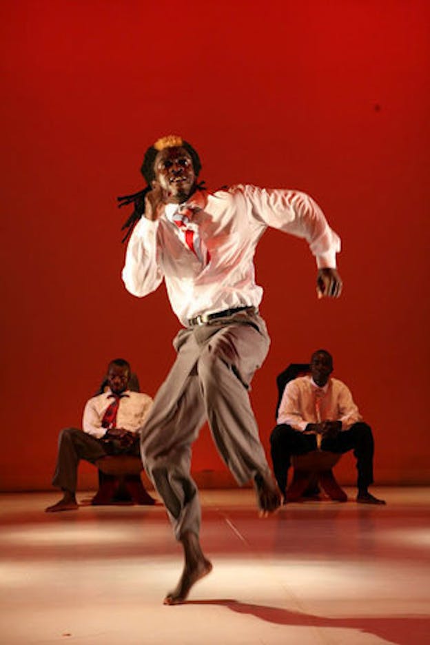 In the center, one performer wearing a tucked white shirt, a patterned necktie, and a pair of fitted pants is in the middle of a leaping motion. In the background, two other performers with similar outfits are seated with open legs. The one on the left sinks into the chair while the one on the right rests their elbow on their knees. The stage has a beige color floor and a red backdrop.
