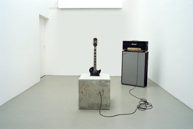 An installation image of a black electric guitar submerged in a concrete cube so only the top half of the guitar is showing. A cable runs from the cube towards an amplifer behind it. The walls of the room are white and the floor is grey. 