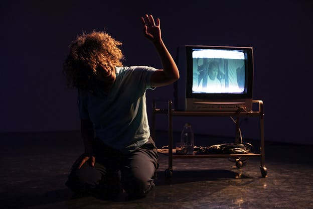 A performer sits with their knees bent besides a television sit. The performer angles their head down, hiding their face from viewers. They are raising their left hand as if about to wave. On the television behind them, a man looks out imploringly besides a religious cross. 