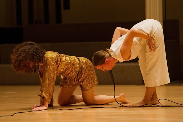 A performer in shiny gold crawls in front of a performer in white bending forwards, a microphone fully immersed in their mouth.