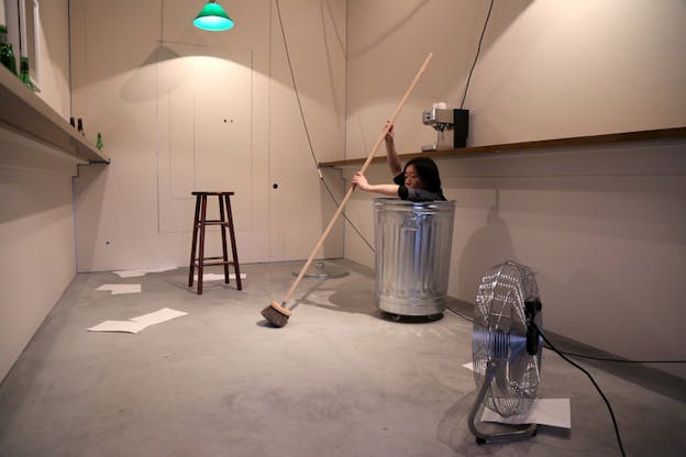 A person inside a metallic garbage bin holds a broom and sweeps the floor, near them farthest from the viewer a stool surrounded by papers on the floor and on the other side closest to the viewer a fan.
