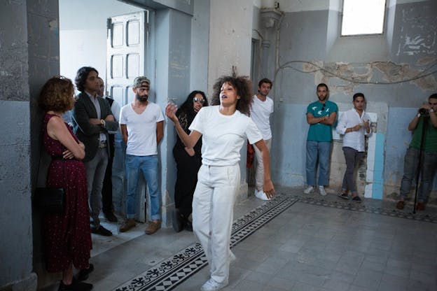 A performer dressed in white swaggers around a room with chipped paint on the walls, as an audience around them watches. 