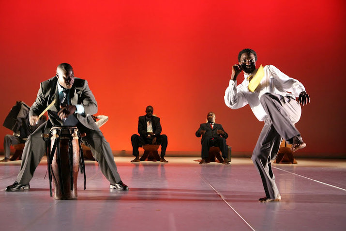 On the left, one performer wearing a grey tuxedo outfit is in a squat position and plays a wooden drum with his hands and a drumstick. On the right, another performer wearing a tucked white shirt, a yellow necktie, and a pair of grey pants leans back and raises his curled left leg and right arm. In the background, three other performers dressed in tuxedo outfits are seated on wooden chairs with tall backs. Two additional chairs beside them are not taken. The stage has a light grey floor and a red backdrop.