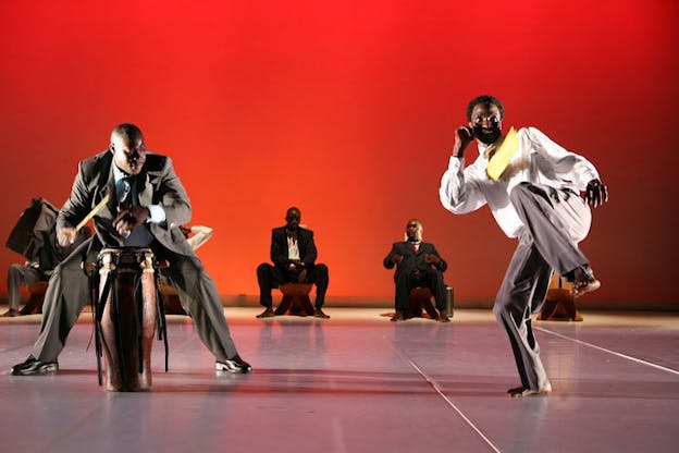 On the left, one performer wearing a grey tuxedo outfit is in a squat position and plays a wooden drum with his hands and a drumstick. On the right, another performer wearing a tucked white shirt, a yellow necktie, and a pair of grey pants leans back and raises his curled left leg and right arm. In the background, three other performers dressed in tuxedo outfits are seated on wooden chairs with tall backs. Two additional chairs beside them are not taken. The stage has a light grey floor and a red backdrop.