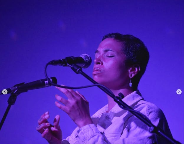 Holland Andrews faces left, bathed in blue light, singing into two microphones with their eyes closed. Both of their hands are slightly raised.