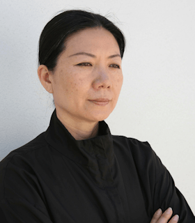 A side angle portrait of Kimsooja in front of a white wall. Her black hair is pulled into a low bun and she wears a black jacket. With her arms crossed in front of her chest, she looks into the distance. 