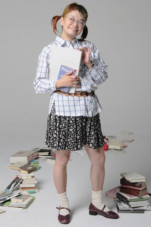 Nexus6 performs in a white space with piles of books on the floor. She wears her hair in pigtail and has wire rimmed glasses, a white shirt with a blue grid on it, a black and white floral skirt, white sock, and red mary jane shoes. She holds two books in her hand, one of which has the title 