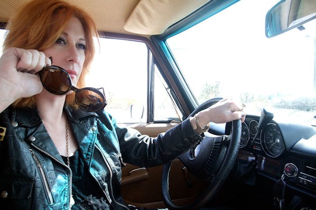 Close-up of a person wearing a black leather jacket sitting in the drivers seat of a car holding the wheel with one hand and lowering their sunglasses with the other, as they look confidently ahead.