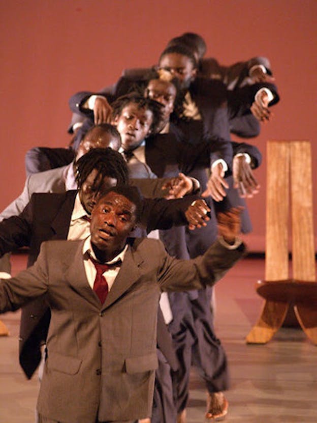 Several performers wearing grey and brown toned tuxedo outfits are lined up in a row. Each of them raises their arms laterally while tilting their heads to the right. A wooden chair with a tall back is placed on the right side of the background. The stage has a caramel colored floor and a brick red backdrop.
