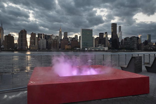 A red box with opening in the middle extracting steam lighted in pink sits on the waterline with high skyscrapers on the other side.