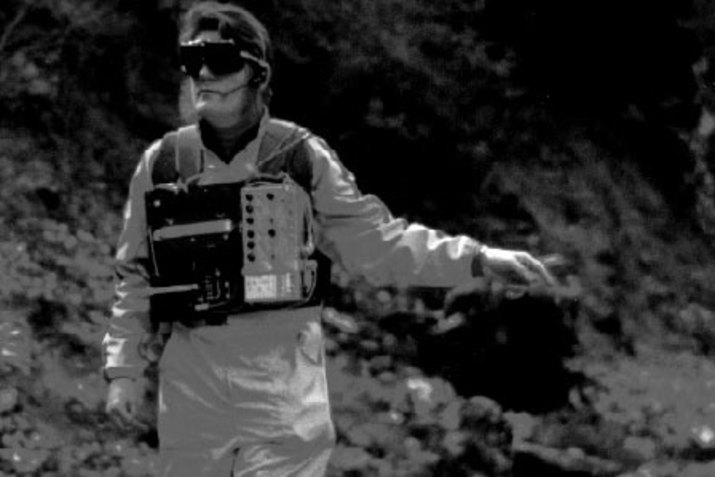 A black and white image of George Mumma wearing a light colored jump suit, a vest of sound equipment and thick sunglasses. He looks directly at the camera and points to his left and down. The background behind him is of blurred foliage. 