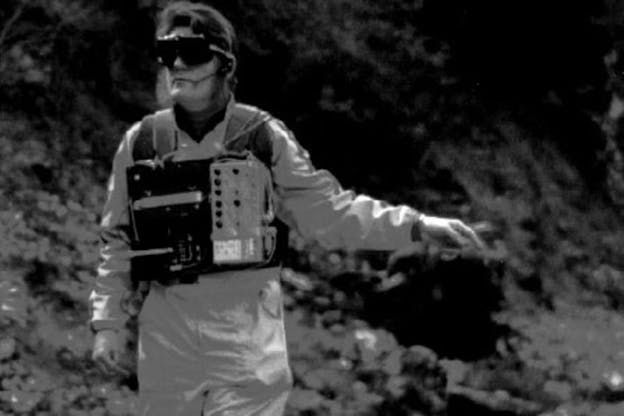 A black and white image of George Mumma wearing a light colored jump suit, a vest of sound equipment and thick sunglasses. He looks directly at the camera and points to his left and down. The background behind him is of blurred foliage. 