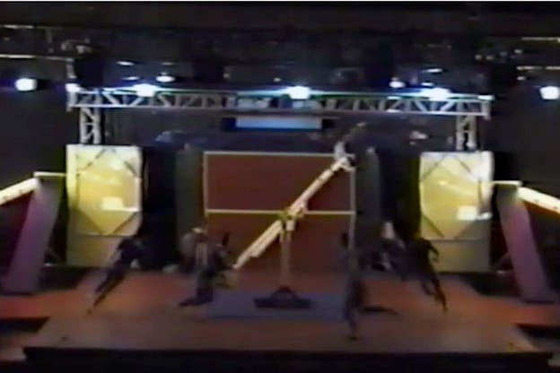 Image of performed clad in blue unitards running in a circle around a yellow custom made gymnastic equipment. 