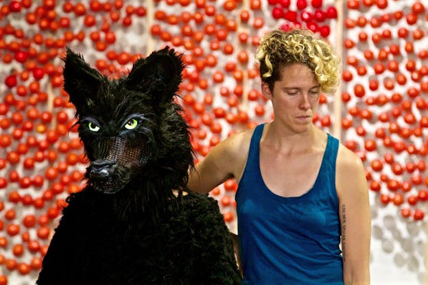 A figure in a blue tank top stands near a person dressed in a black wolf costume in front of a wall with red spheres.