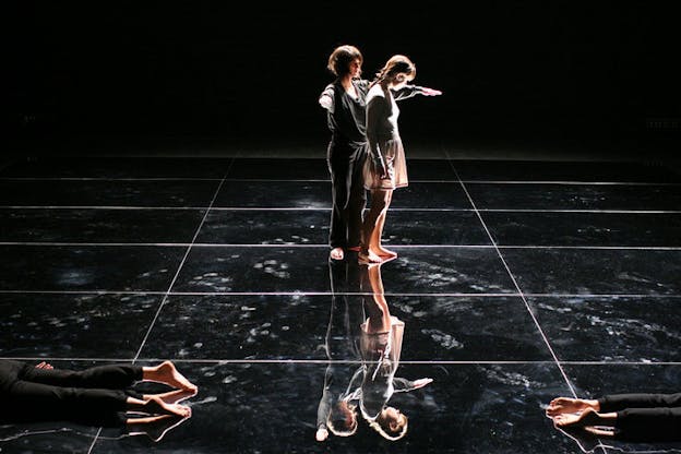 On a black-lit reflective stage, a performer raises their arms in an L-shape around the bent shoulder of another performer. On each of the front-sides, bare feet lying on the floor are visible.