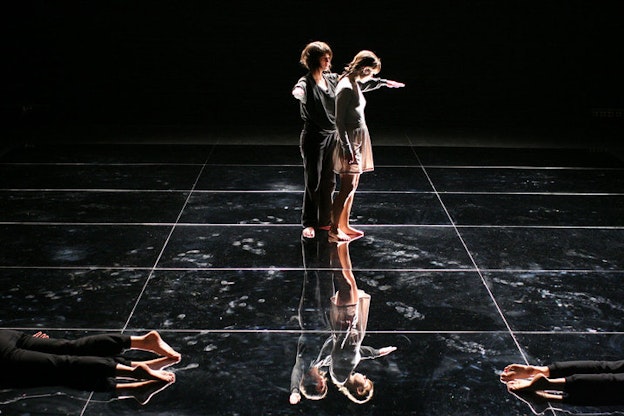On a black-lit reflective stage, a performer raises their arms in an L-shape around the bent shoulder of another performer. On each of the front-sides, bare feet lying on the floor are visible.
