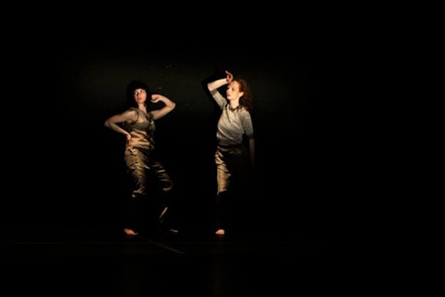 Two persons standing in pitch black stage, their bodies slightly illuminated, strike poses by bending an arm towards their face and looking sideways.