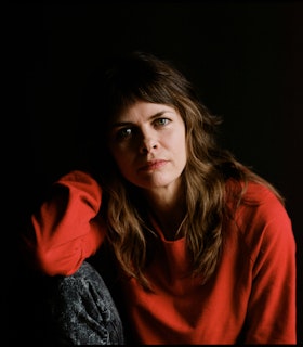 Joanna Kotze sits before a stark black backdrop and looks directly at the camera, the right side of her face is shadowed. Her right knee is raised and her right elbow rests on it. She is dressed in a bright red sweater and gray acid-wash jeans. 