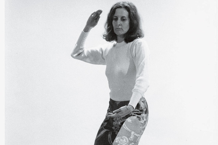 Black and white photograph of dancer in a sweater and patterned jeans bending their knees slightly and raising one arm in a knifehand strike position and making a downward oval shape with their other.