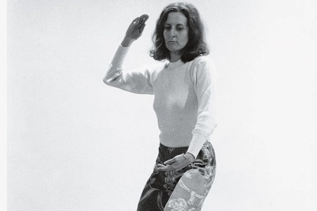 Black and white photograph of dancer in a sweater and patterned jeans bending their knees slightly and raising one arm in a knifehand strike position and making a downward oval shape with their other.