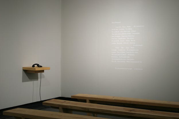 An exhibition photograph of a gray wall with a poem written on it in white letters. There are three wooden benches in front of the wall and a wooden shelf with a black telephone on it.