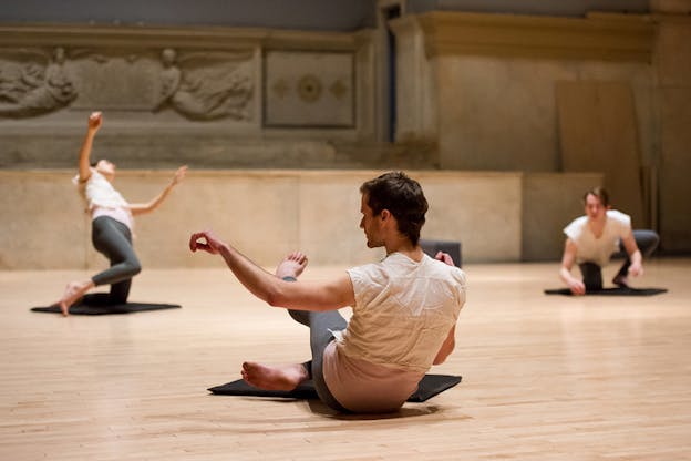 Three performers on small black mats in various poses. The one on the far left supports their body on one knee, while the one on the right crouches with one knee. The person in the middle closest to the viewer stands with their back to us, seated down with their arms and one leg mid-air. 