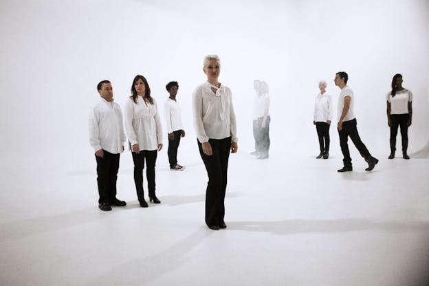 A group of people dressed in white shirts and black pants positioned throughout a blank white room turning their heads to look forwards with the exception of one person walking in profile, while two persons in the background are slightly obscured by a foggy white substance.