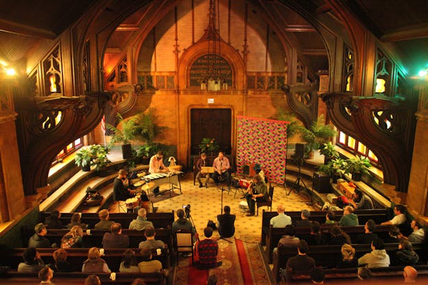 Performers behind musical instruments sit at the apse of a chapel as a seated audience faces them from the pew. Behind the musicians various pots with green leaves, a tapestry with colors of fuchsia, green and blue sits in between the plants on the right side. 