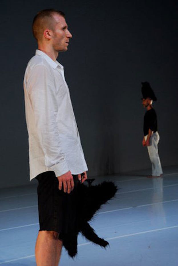 A performer in the foreground of a sparse, blue hued room, stands looking forlorn and holding a fuzzy black object. In the background, another performer stands, facing the opposite direction and wearing the fuzzy black object atop their head. 