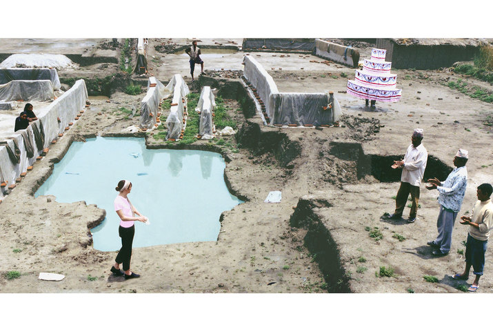 A group of people on a construction site wiping their hands. On the upper right corner, one person is dressed in a four tier cake costume.