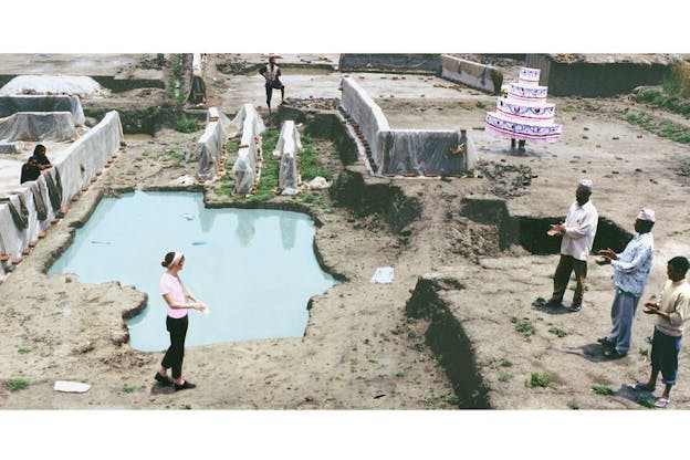 A group of people on a construction site wiping their hands. On the upper right corner, one person is dressed in a four tier cake costume.