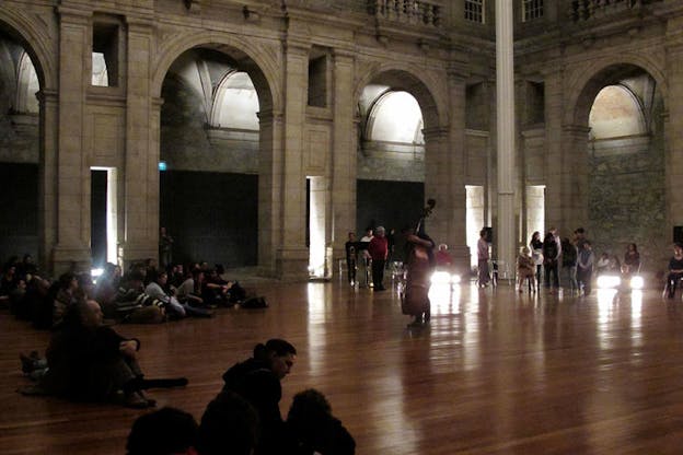 Viewers gather on the floor watching a double bassist perform in a dimly lit spacious room enclosed by tall stone arches. 