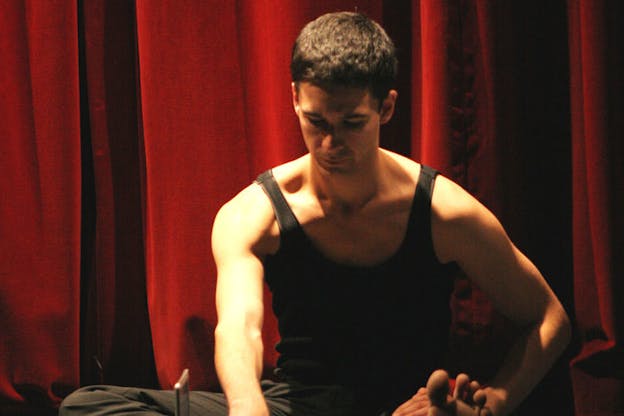 Jonah Bokaer performs in front of a red curtain. He wears a black tank top and black pants and is barefoot. He sits on the floor and bends over his extended left leg. 