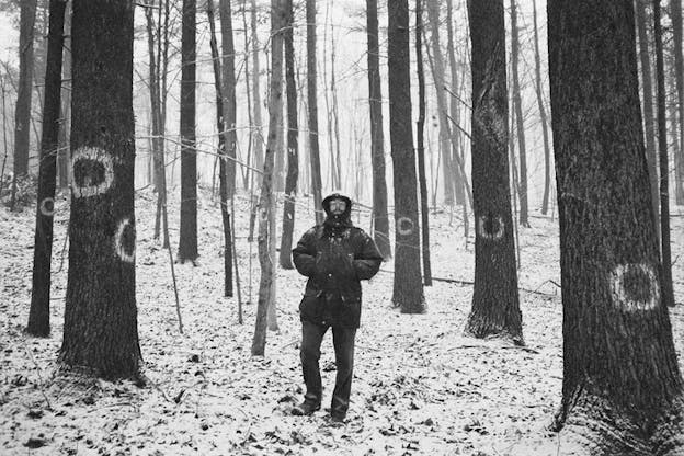Black and white photograph of Barr dressed in a snowcoat with his hands in his sleeves, standing in a snowy forest of thin trees with white circles painted on their dark trunks. 