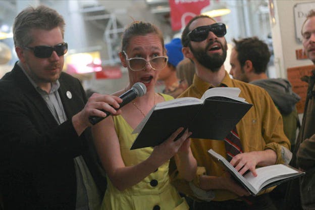 A person dressed in a yellow dress and glasses holding a book and talks in a microphone that a person next to their side is holding.
